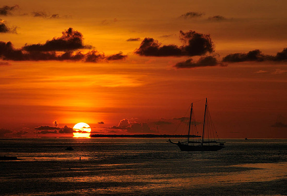Dark orange and red skies, with bright yellow and white sun melting into horizon. Schooner in foreground.